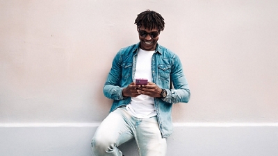 A man in a white t-shirt, denim jacket, jeans, and sunglasses smiles as he looks down at his phone. 