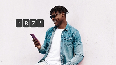 A man in a white t-shirt, denim jacket, and sunglasses smiles at his phone as he uses it. 