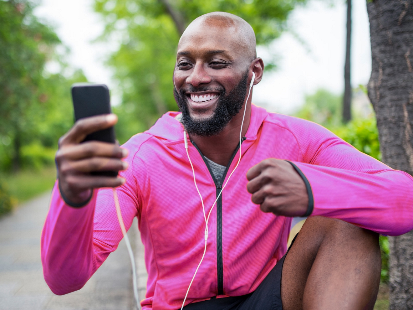 A man in a magenta jacket and wired earbuds sits in a park and smiles at his phone.