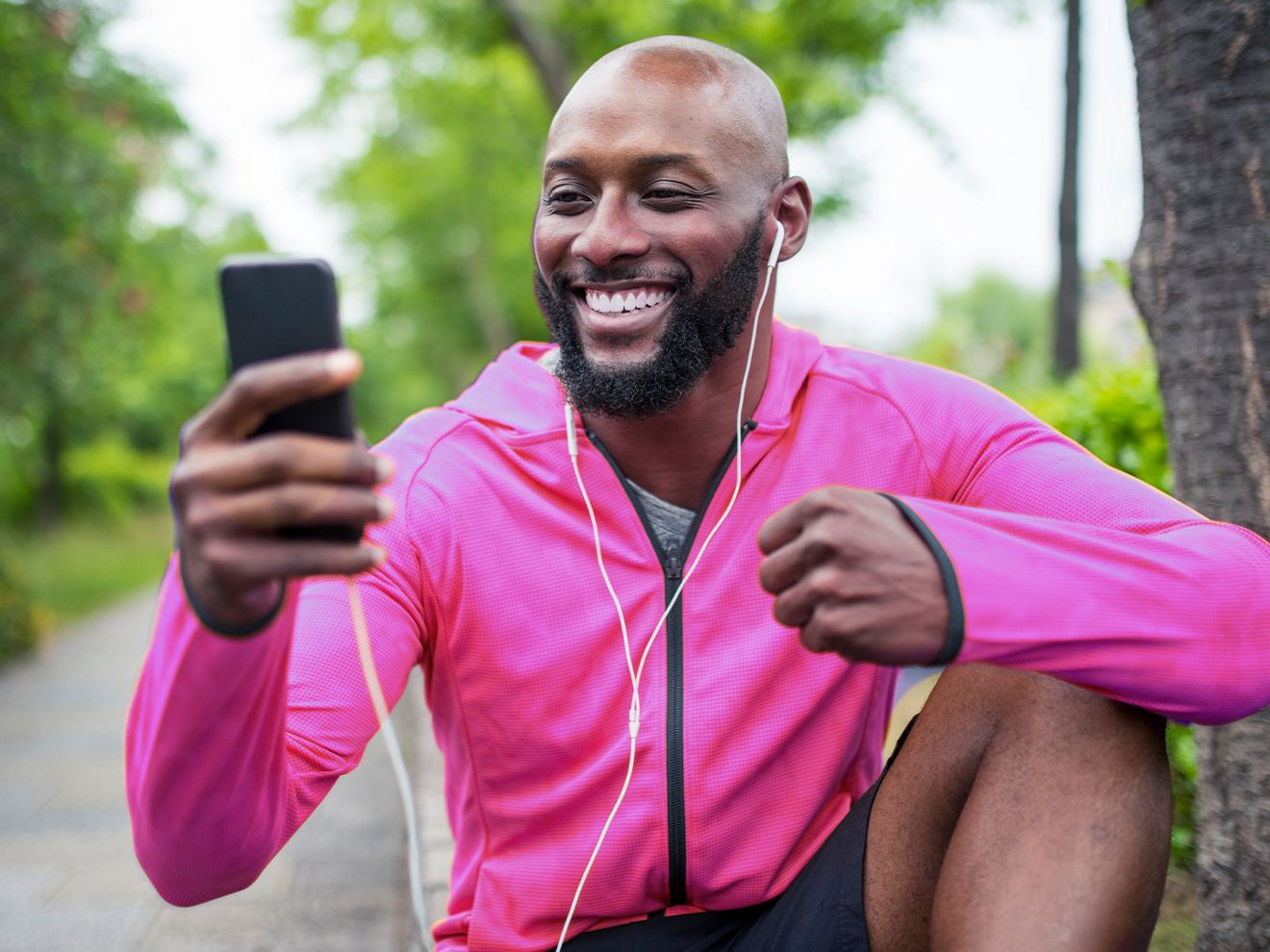 A man in a magenta jacket and wired earbuds sits in a park and smiles at his phone.