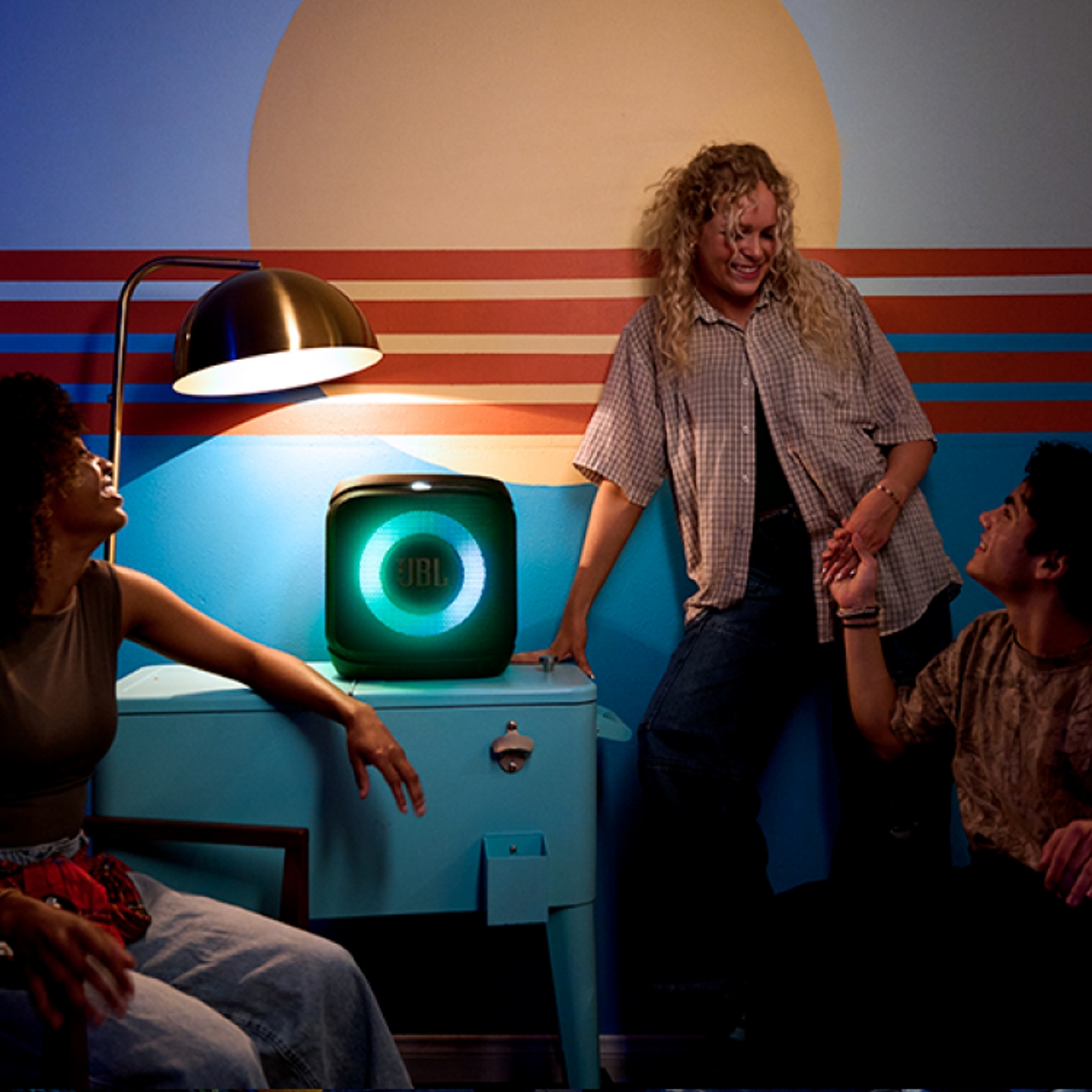 A JBL speaker glowing blue while three friends hang out in a dimly-lit room.