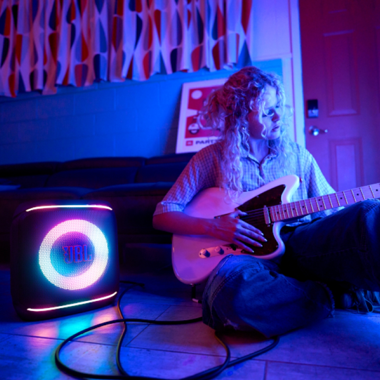 A young woman playing an electric guitar in a dimly-lit room while sitting next to a glowing JBL speaker.