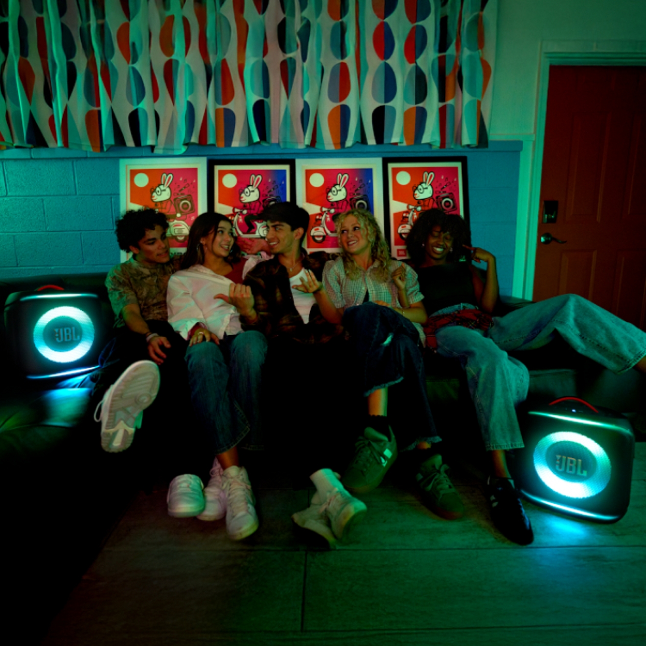 Two glowing JBL speakers on the floor while five friends hang out on a couch.