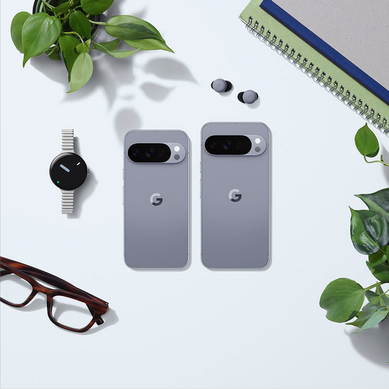To compare sizes, a Moonstone Google Pixel 10 Pro and Moonstone Google Pixel 10 Pro XL lay side by side screen side down on a flat surface surrounded by plants, glasses, notebooks, earbuds, and a watch. 
