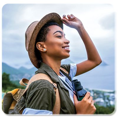 A smiling woman tips her hat back as she looks at a surrounding mountain and sea view.