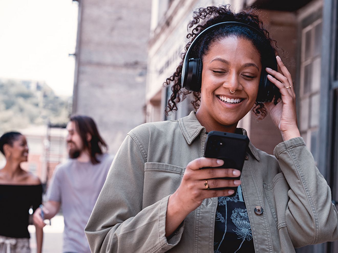 A woman wearing headphones looks at her phone and smiles on a city street, with blurred pedestrians behind her.