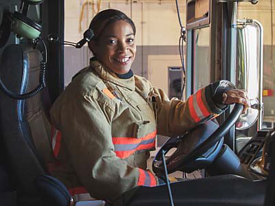 A firefighter smiles as she sits behind the wheel of a fire truck.