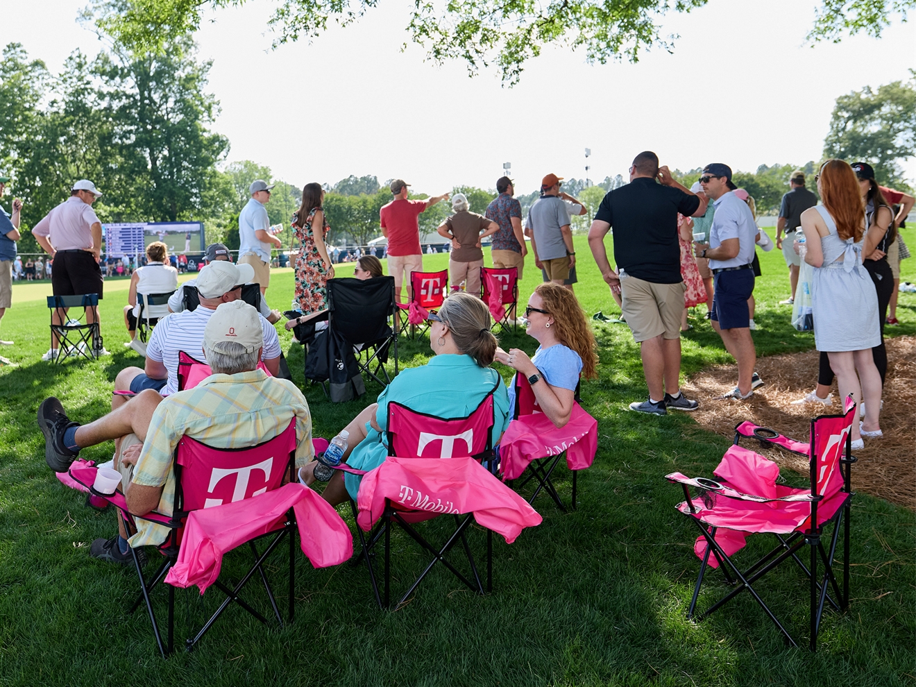 A group of people gathered on the edge of a golf course. Some are standing and some are sitting in T-Mobile branded lawn chairs. 