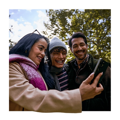 A woman in a magenta and white plaid scarf stands outside, surrounded by a couple of men. All of them smiling at a phone.