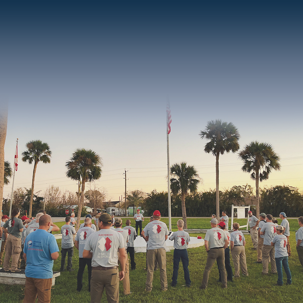 In a park lined with palm trees, members of Team Rubicon listen to instructions from a team leader. 
