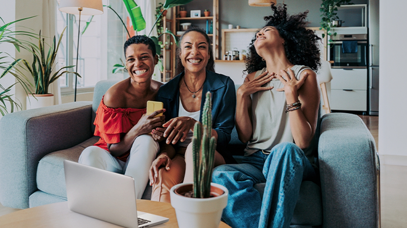 Three women smiling on a couch with a laptop open in front of them.