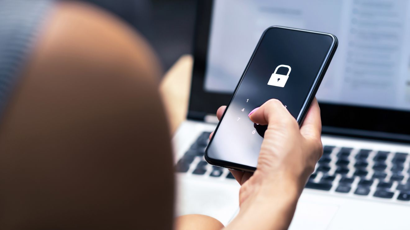 Close-up of hands holding a smartphone with a lock icon overlay and a laptop in background.