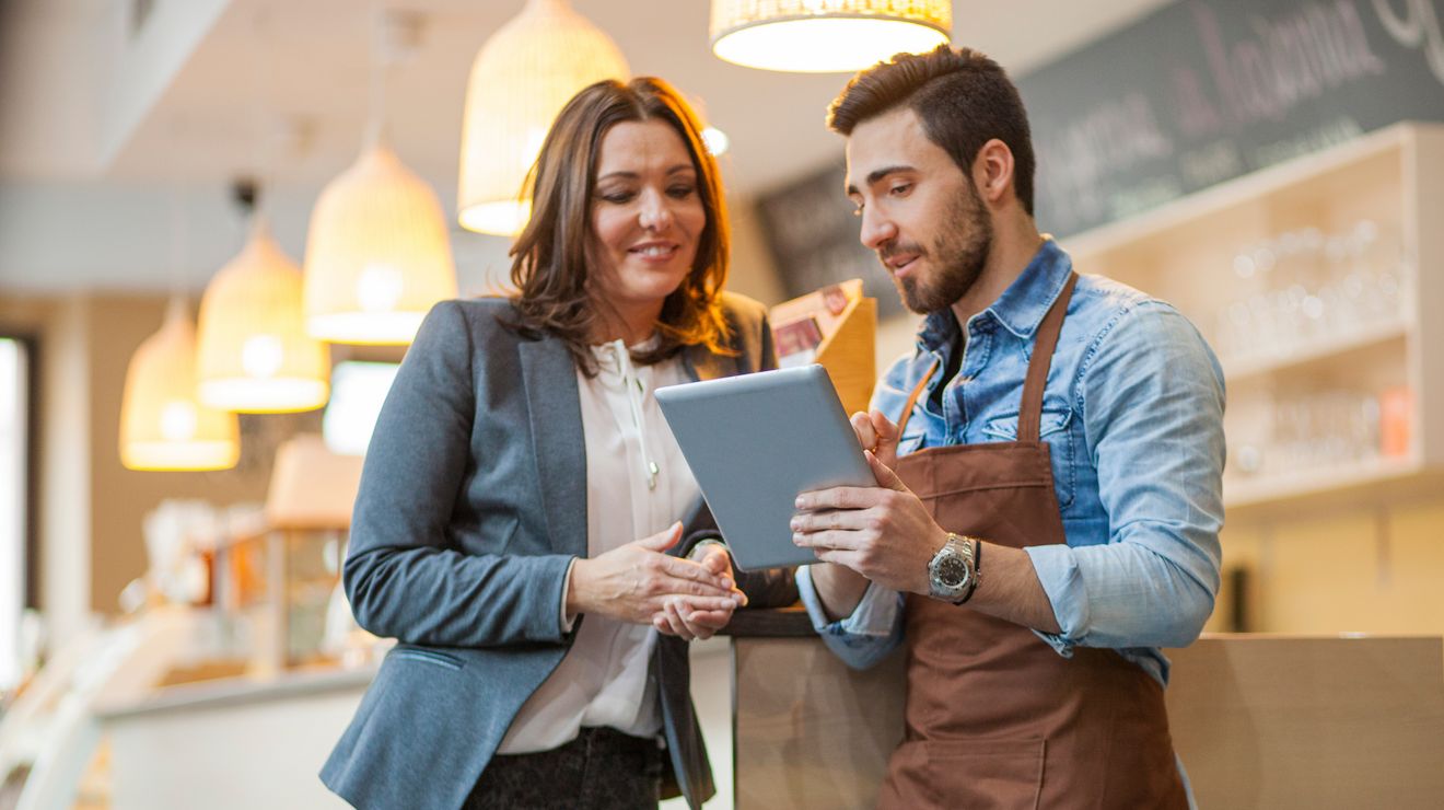 A manager and barista use a tablet to review retail operations in a coffee shop.