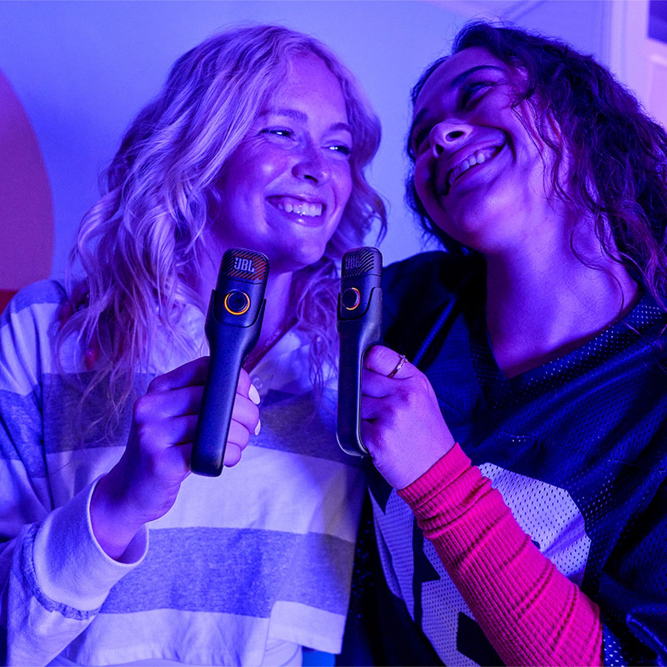 Two women smile at each other while holding black JBL EasySing wireless microphones with illuminated control rings under purple and blue lighting.