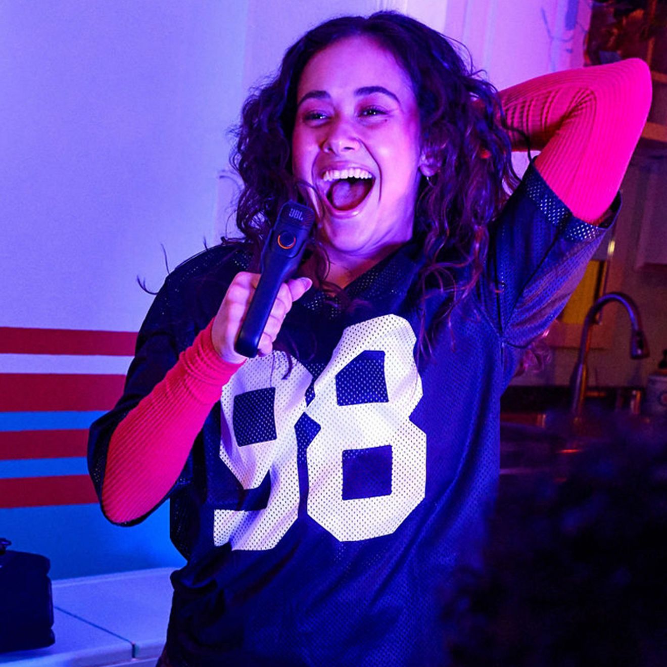 A woman sings into a black JBL EasySing wireless microphone, smiling with one arm raised behind her head under vibrant purple and pink lighting.