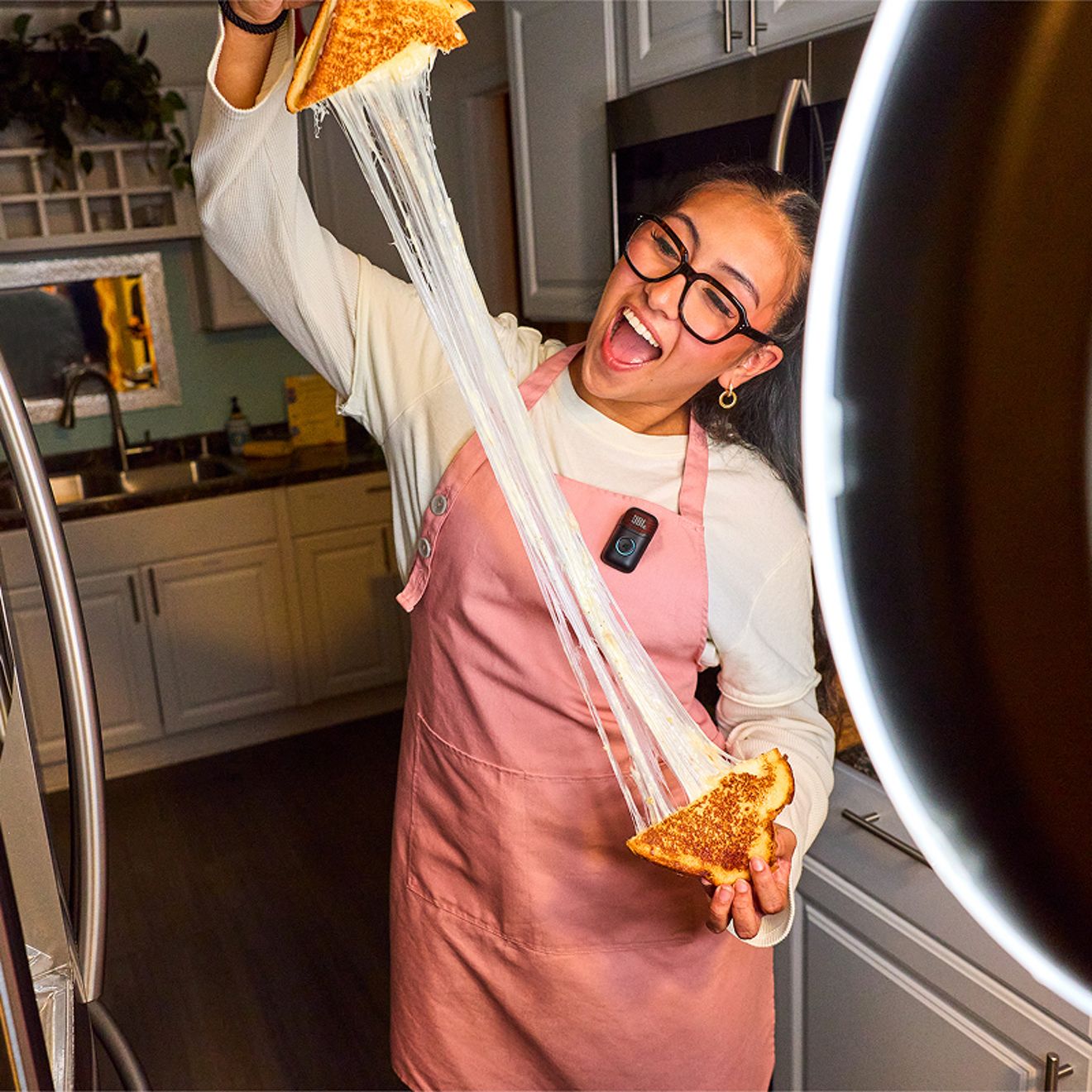 A woman wearing a JBL EasySing Mic Mini clipped to her pink apron stretches melted cheese from a sandwich while filming herself with a ring light in a kitchen.