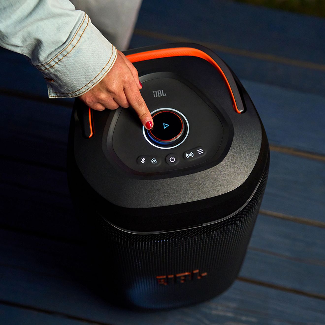 A hand presses the illuminated play button on top of a black JBL PartyBox 130 speaker with orange accent lighting.