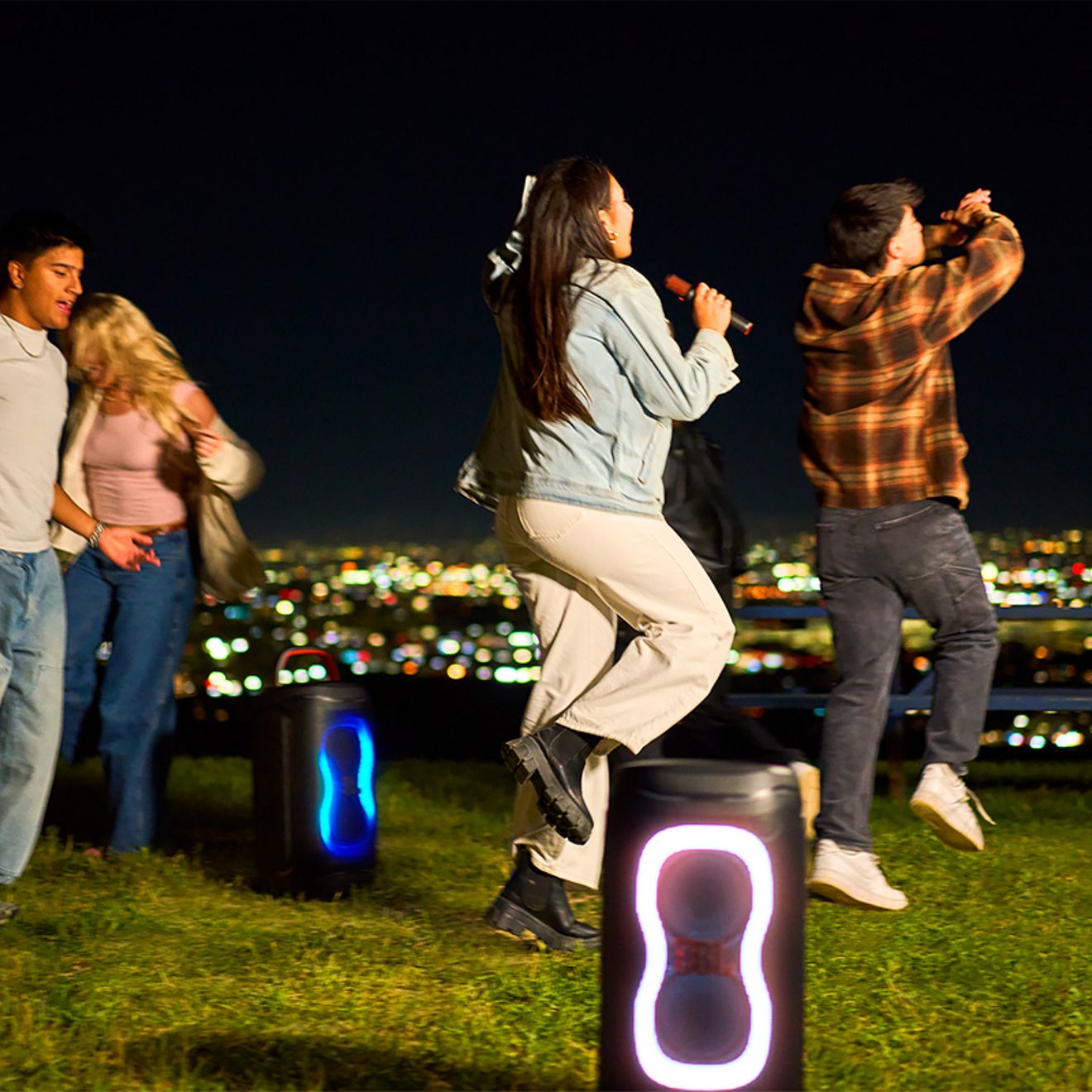 Two JBL PartyBox 130 speakers with colorful LED lights play as a group of friends dance and sing with a microphone outdoors at night, overlooking a city skyline.