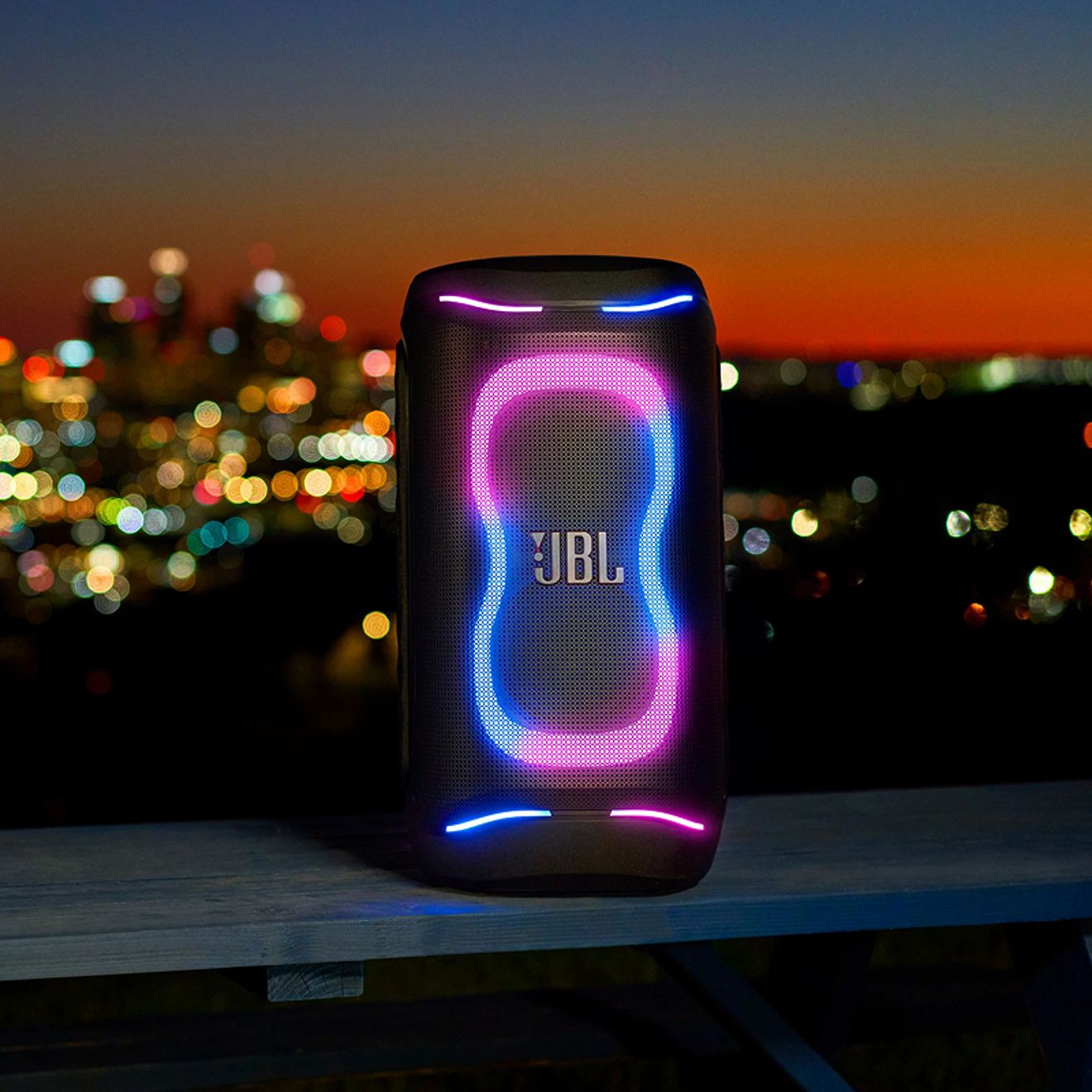 A black JBL PartyBox 130 speaker displays a vibrant blue and pink LED lightshow against a blurred city skyline at night.