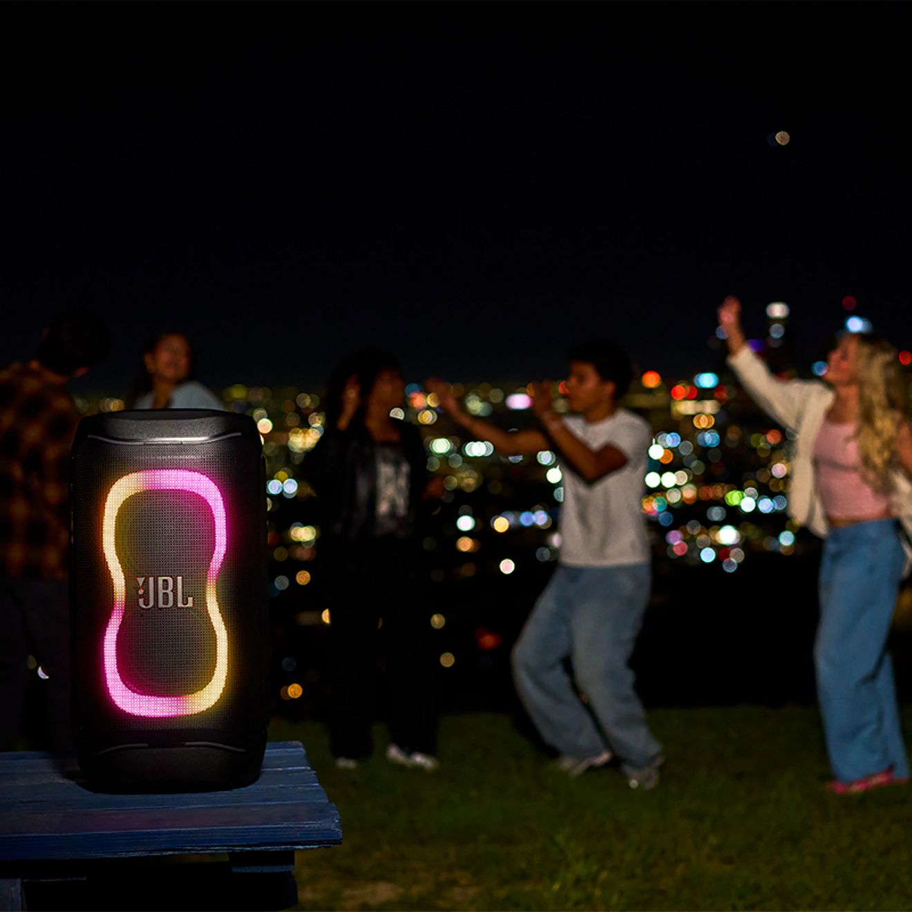 A JBL PartyBox 130 speaker with colorful LED lights plays at an outdoor nighttime party as a group of friends dance against a glowing city skyline.