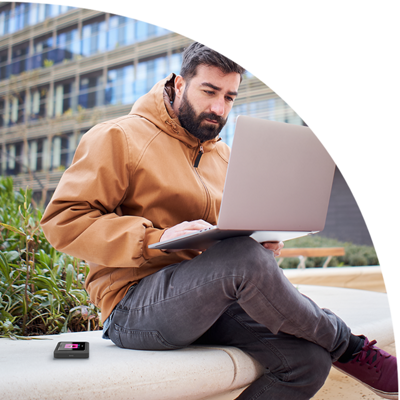 Man with laptop and hotspot device sitting outside near a tall building.