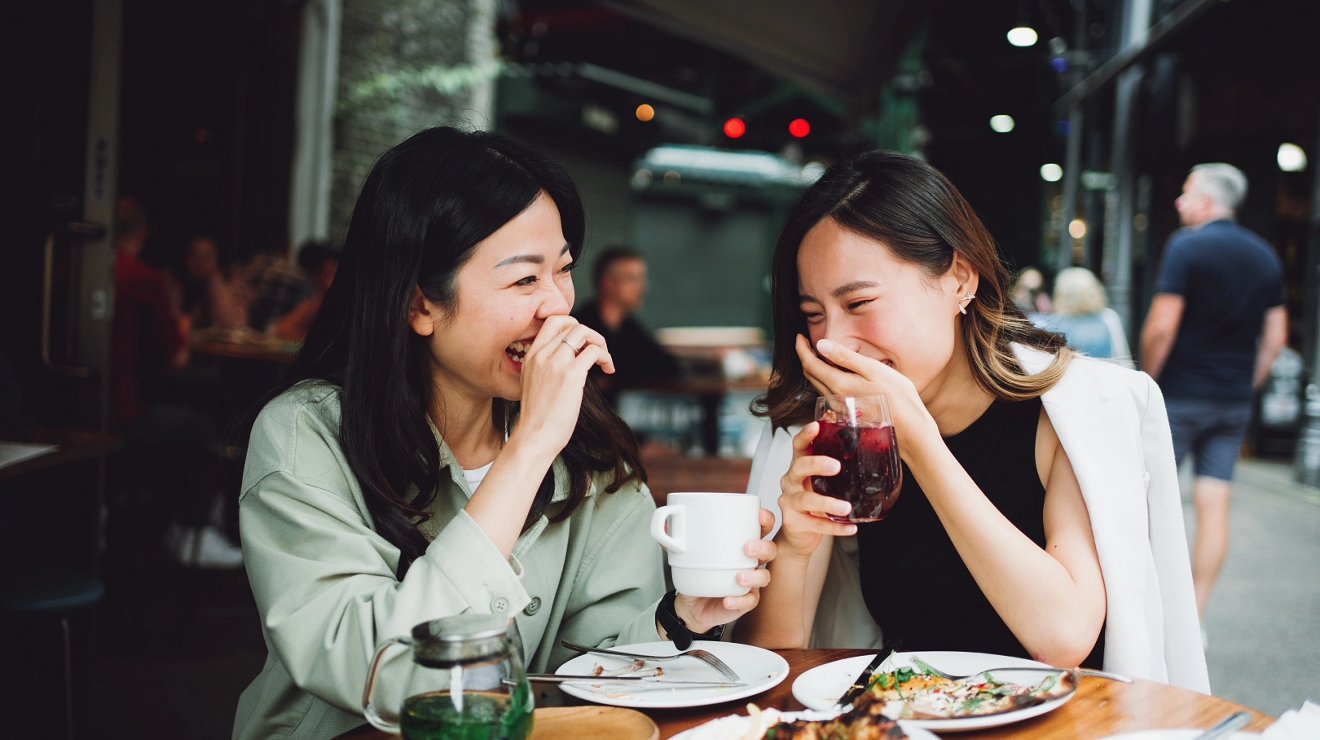 Two people laughing and enjoying a meal on a patio in a city