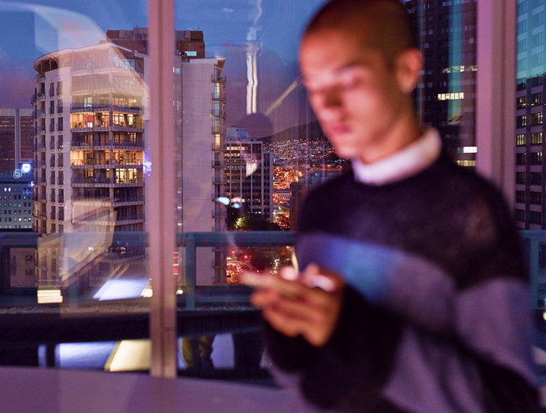 A person stands by a window in their city apartment and scrolls through their phone messages.