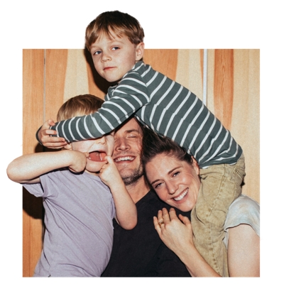 A young boy sits on his smiling mother’s shoulders and puts his arms around his sibling in his dad’s arms, who is making a goofy face. 