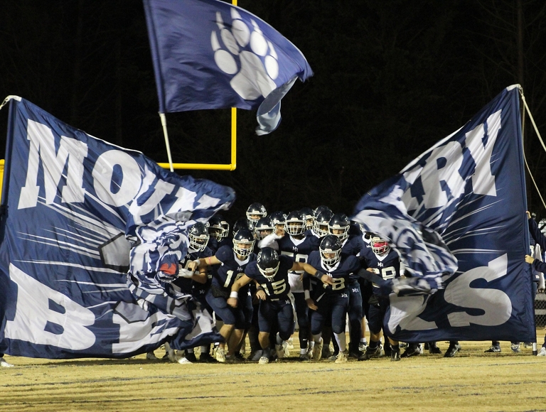 The Mount Airy football team, wearing navy and white uniforms, runs through a banner onto the field.