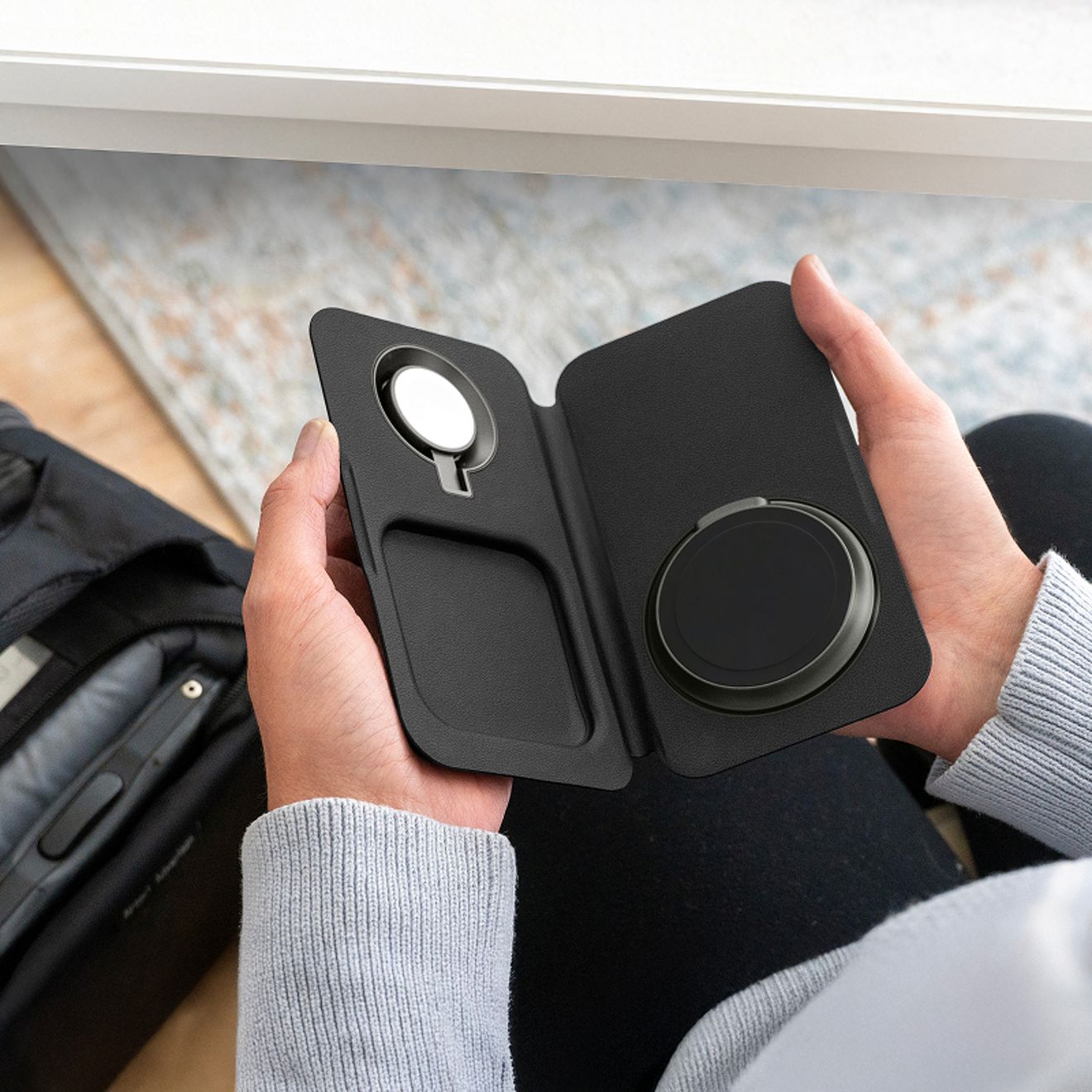 A person holds an unfolded black Nimble Fold 3-in-1 wireless travel charger, showing dedicated charging areas for a smartphone, Apple Watch, and wireless earbuds.