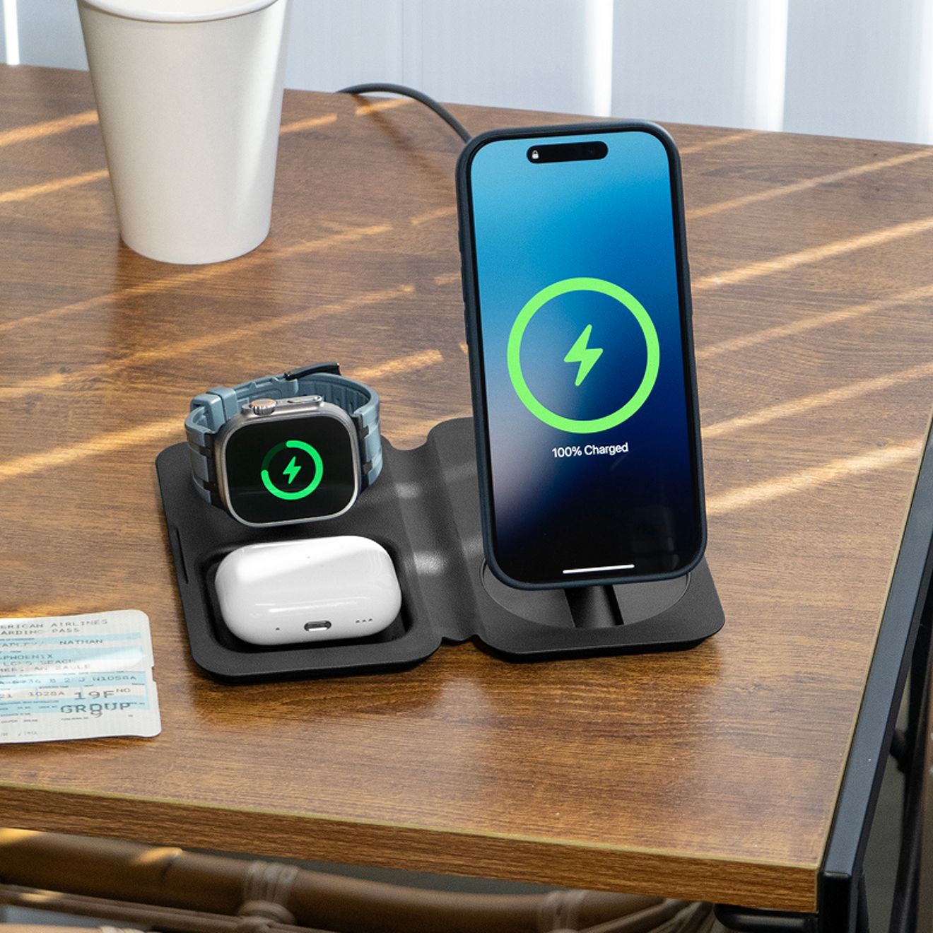 A smartphone, Apple Watch, and wireless earbud case charge simultaneously on an unfolded black Nimble Fold 3-in-1 wireless travel charger on a wooden table.