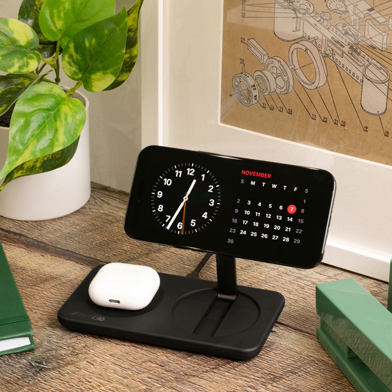 A smartphone and white wireless earbud case charge simultaneously on a black Nimble 2-in-1 magnetic wireless charging stand on a wooden bedside table.