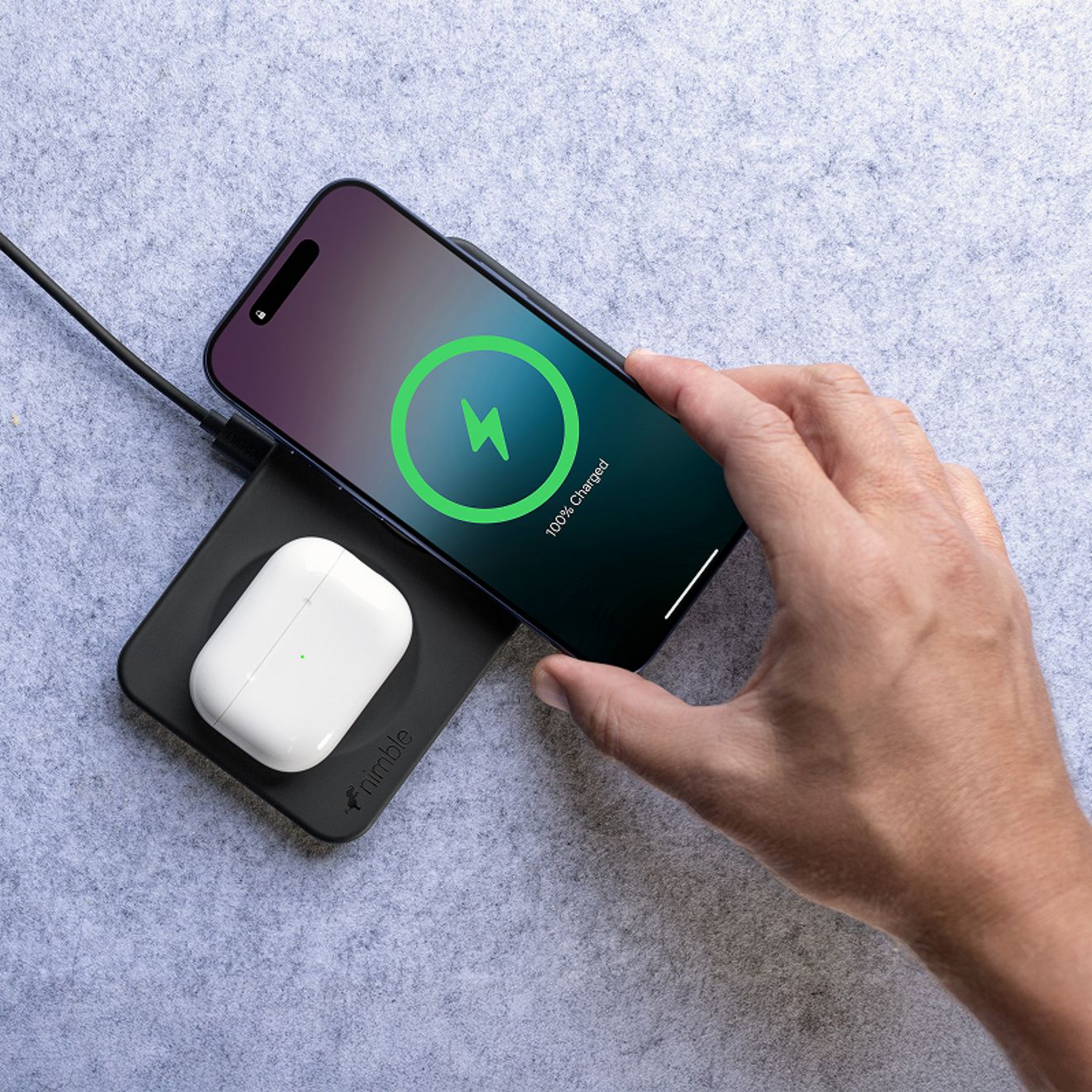 A hand lifts a smartphone showing a full charge from a black Nimble Podium 2-in-1 wireless charging dock while a white wireless earbud case charges on the base.