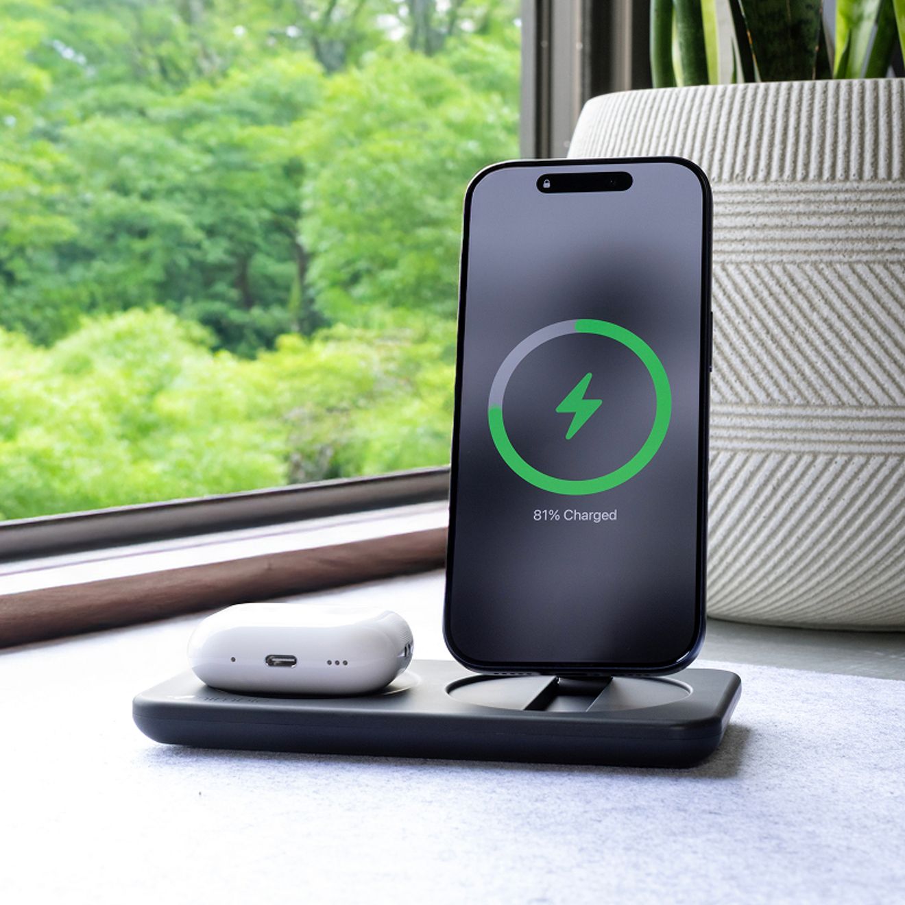 A smartphone and white wireless earbud case charge simultaneously on a black Nimble wireless charging dock beside a window.