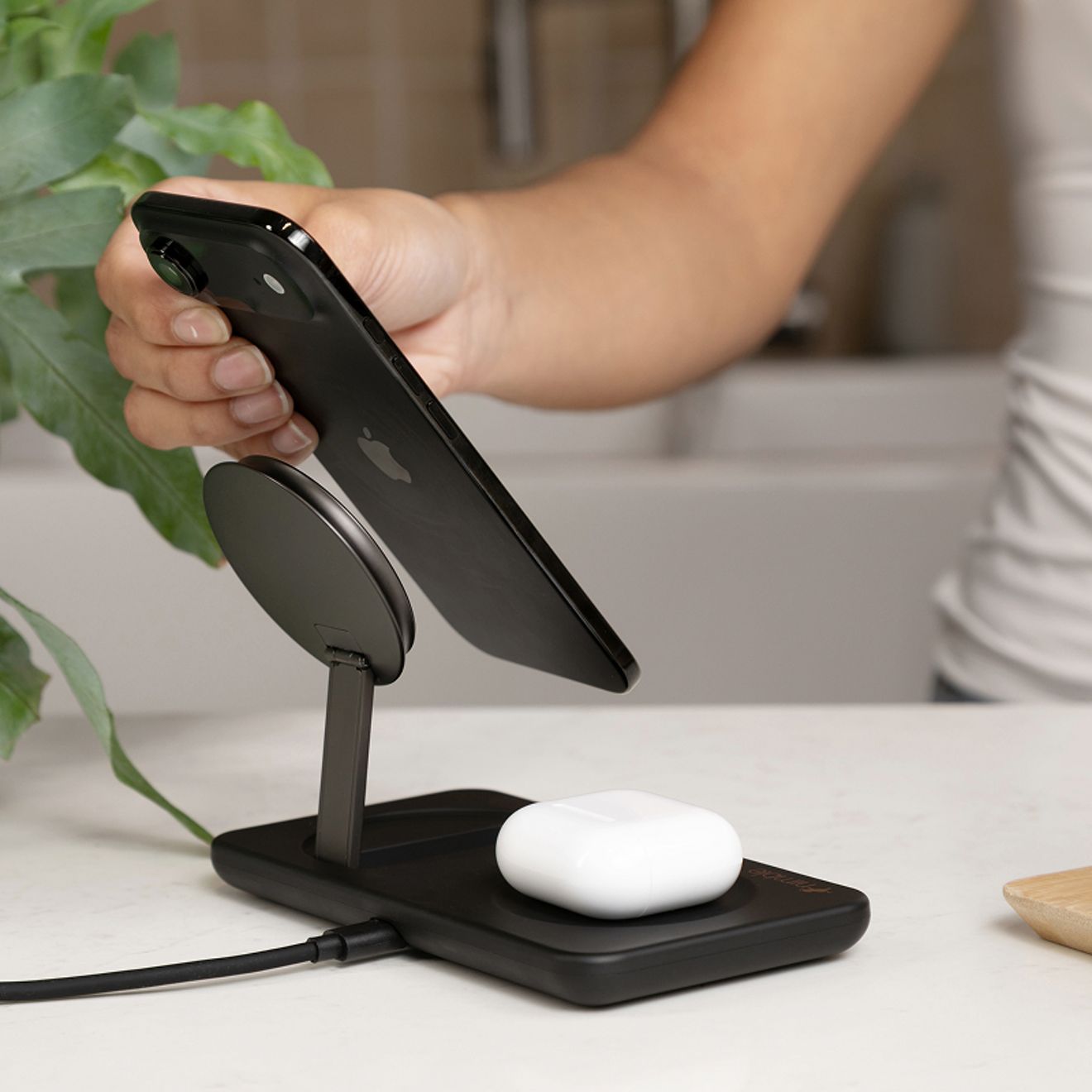 A person lifts a smartphone from a black Nimble 2-in-1 magnetic wireless charging stand while a white wireless earbud case charges on the base.