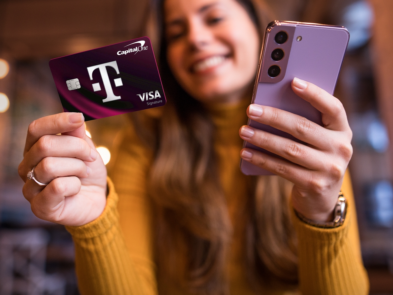 A woman smiles as she holds the T-Mobile Visa in one hand and a cell phone in the other. 