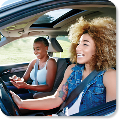 A smiling woman drives while another woman types on a phone.