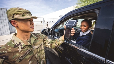 A guard in fatigues uses a mobile device to clear a driver at a facility gate.