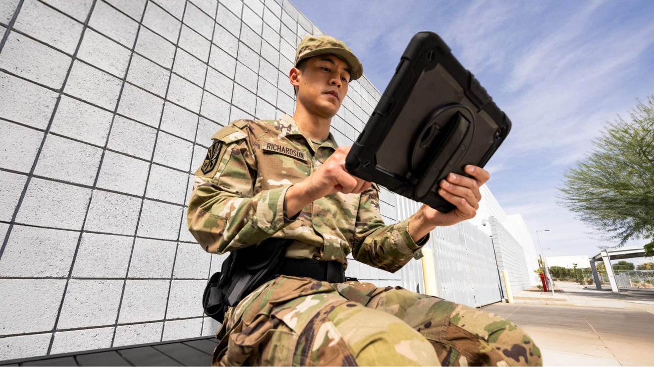 A uniformed guard at a facility gate checks data on a connected tablet.