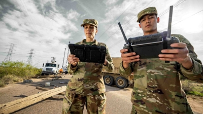 Two personnel in camouflage fatigues hold a connected tablet and a drone controller. 