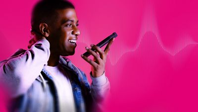 A man in a white t-shirt and denim jacket smiles as he holds a phone up to speak into it.