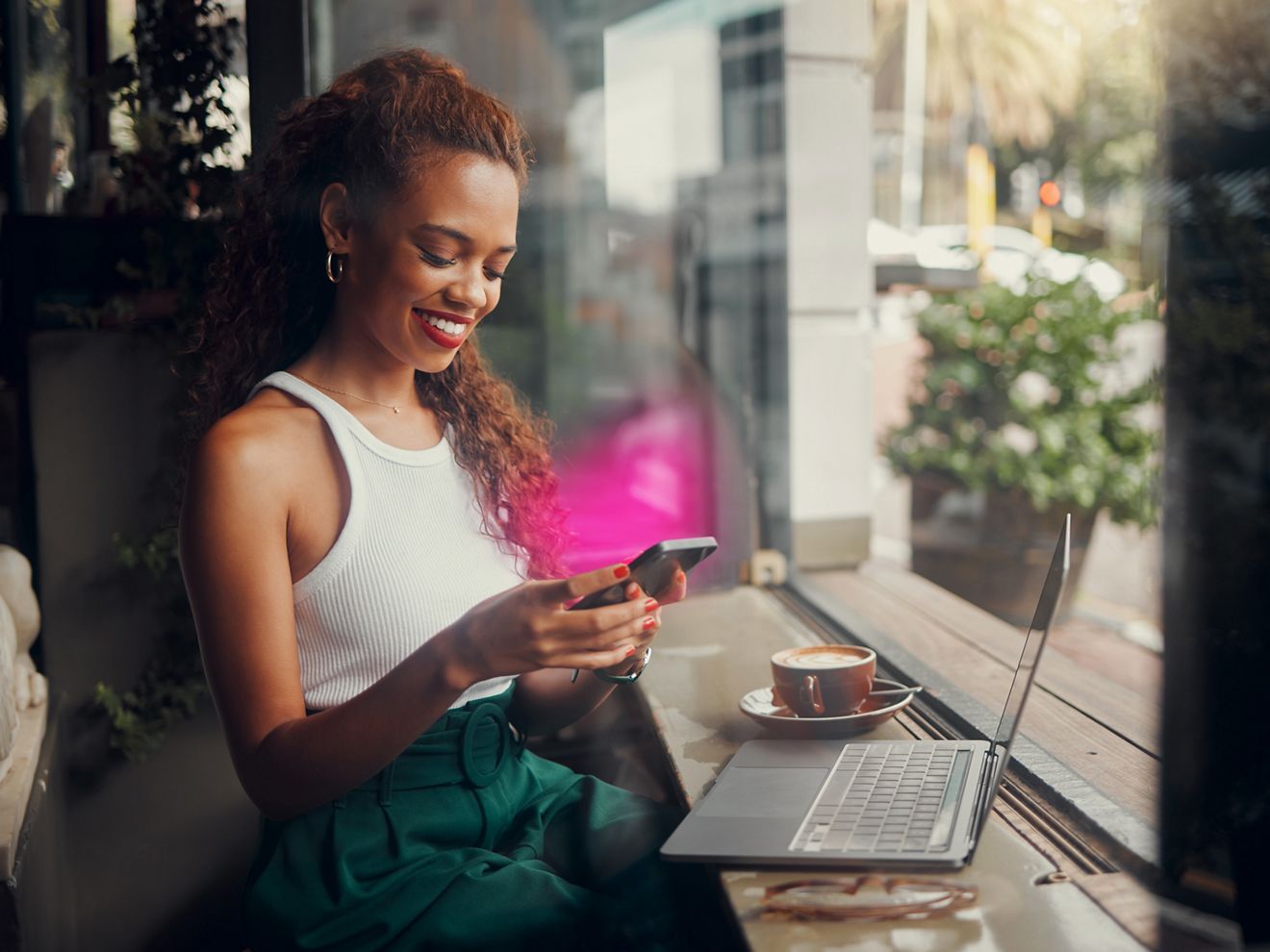 Woman using her phone and laptop along with a cup of copy. Phone has a T-Mobile magenta glow.