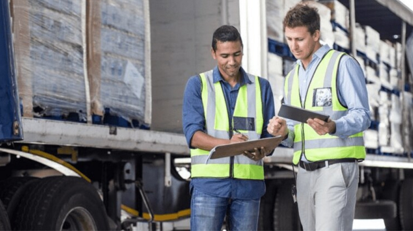 Two freight workers compare the contents on a clipboard to the contents on a tablet.
