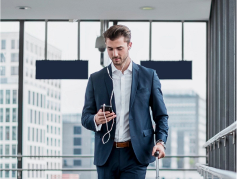 A young professional uses his earphones and looks at his phone while carrying his suitcase through an airport