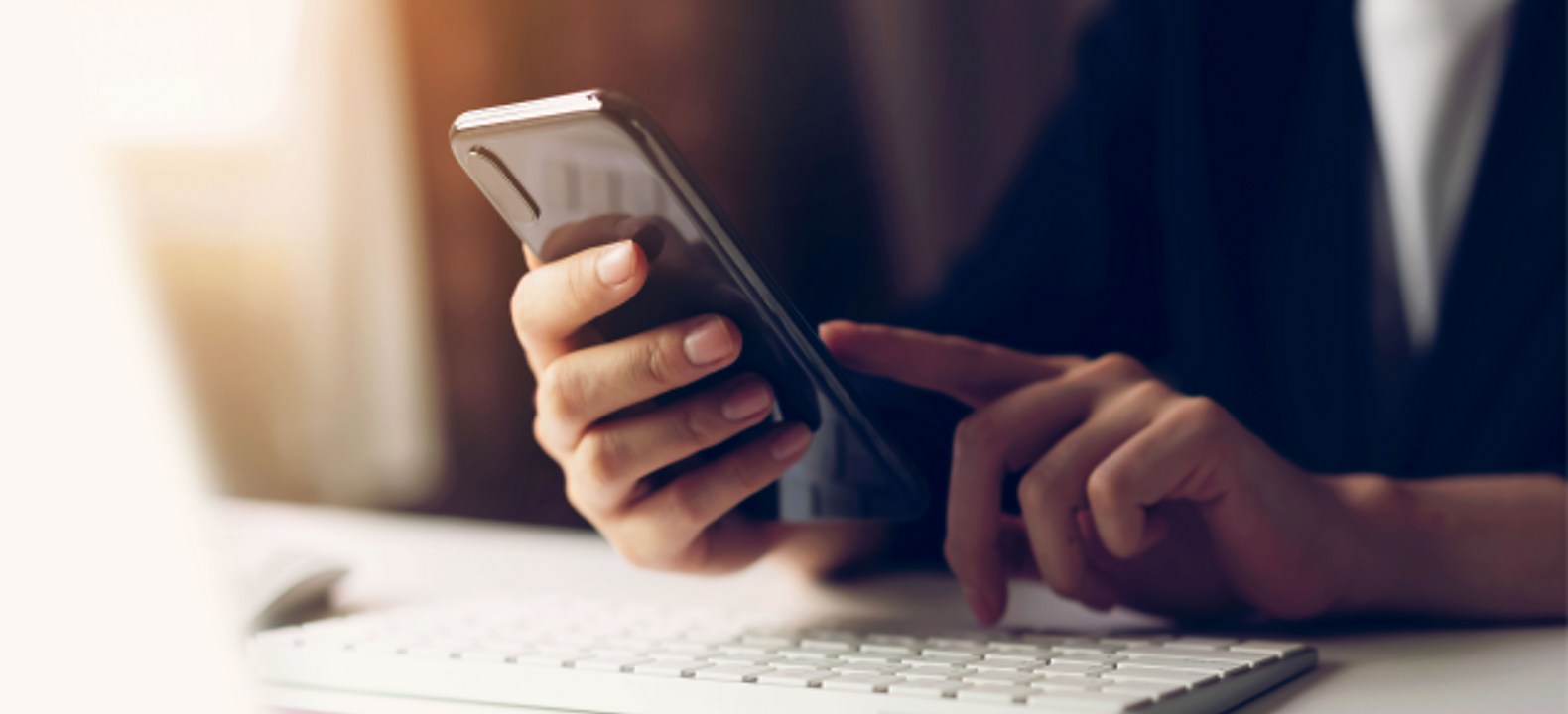 Close-up of two hands using a smartphone above a computer keyboard.