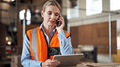 A transportation and logistics professional with a safety vest makes a phone call and views a tablet.
