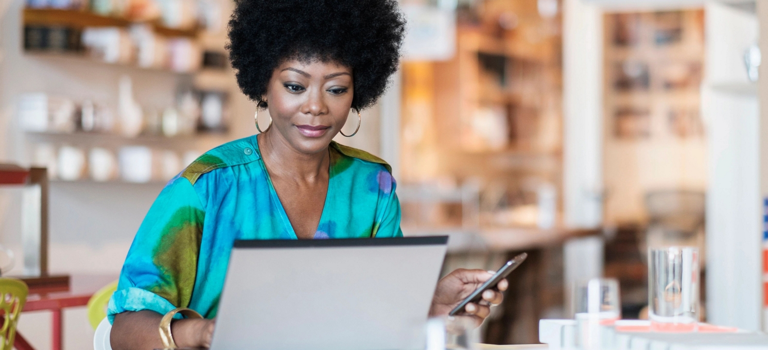 A professional works on her computer while holding a smartphone.