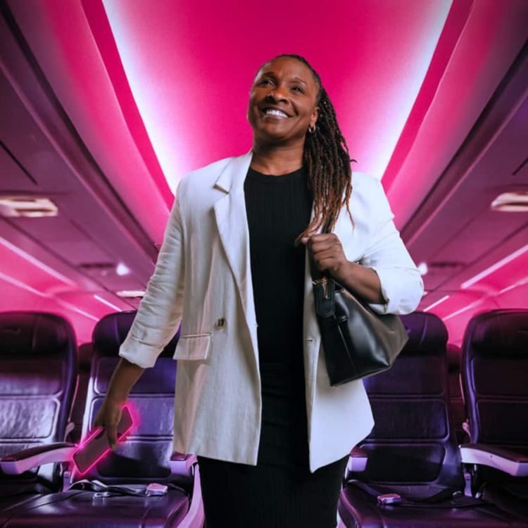 A smiling business traveler walks down a magenta-lit airplane aisle, smartphone in hand.