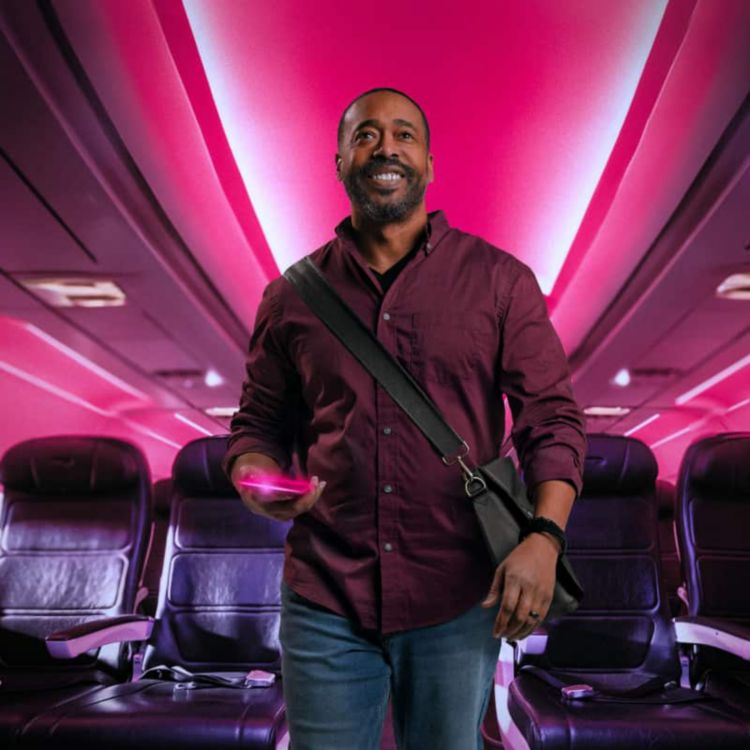 A smiling business traveler walks down a magenta-lit airplane aisle, smartphone in hand.
