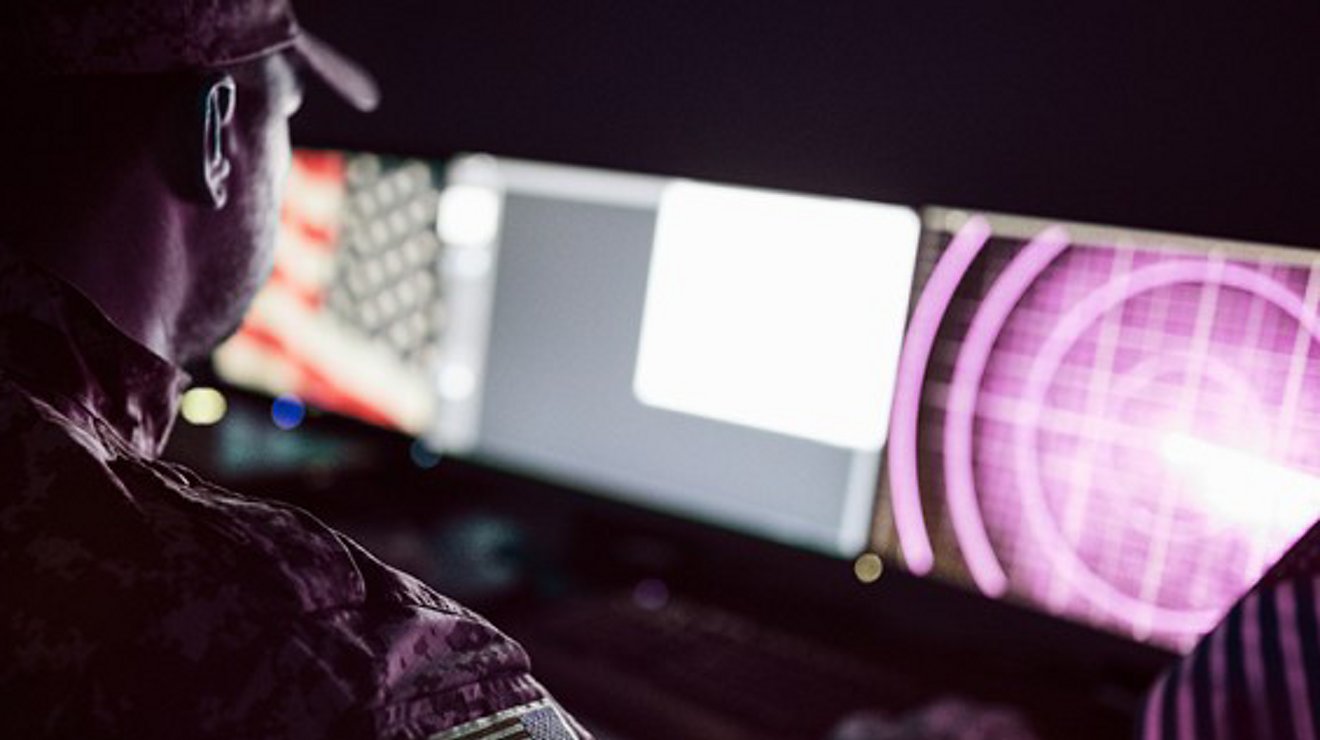 A person dressed in camouflage attire monitors multiple screens in a dark control room.